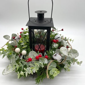 May include: A black metal lantern with a handle sits in the center of a circular wreath. The wreath features green foliage, frosted eucalyptus leaves, small red berries, white berries, and white jingle bells. A red candle is visible inside the lantern.