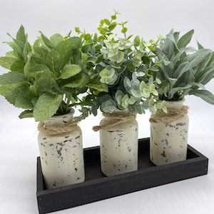 May include: Three decorative vases with faux greenery sit on a dark wooden tray. The vases are off-white with a distressed finish and are wrapped with twine. The greenery includes various shades of green leaves, creating a natural aesthetic.