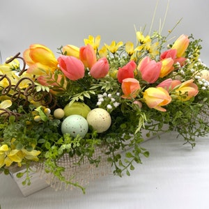 May include: A rectangular wooden planter filled with colourful spring flowers and speckled Easter eggs. The arrangement features yellow and pink tulips, green foliage, and decorative eggs, creating a festive Easter centrepiece.