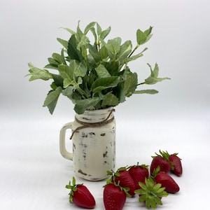 May include: A white distressed mason jar with a handle and twine wrap, filled with green leafy stems. Several artificial red strawberries with green stems are arranged on the surface in front of the jar, creating a decorative display.