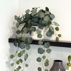 May include: A trailing artificial eucalyptus plant with rounded, sage green leaves cascading from a white ceramic pot featuring a textured, dotted surface. The plant is positioned on a black shelf, displaying multiple long, thin stems.