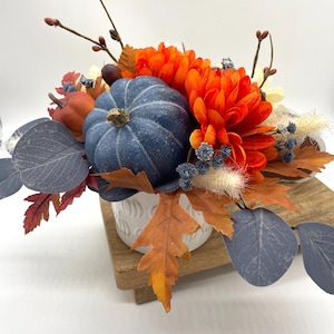 May include: A fall-themed floral arrangement featuring a blue pumpkin as the centerpiece, surrounded by orange chrysanthemums, fall leaves, and blue-gray eucalyptus leaves. The arrangement is in a white, textured vase, set on a wooden base.