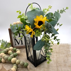 Puede incluir: Linterna de metal negro con asa, llena de girasoles amarillos y follaje verde. Un letrero de madera con las letras "HOME" es visible en el fondo, junto con una guirnalda de cuentas de madera. La linterna tiene una vela.