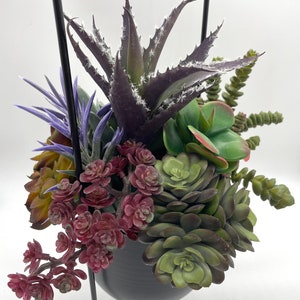 May include: A collection of artificial succulents in various shades of green, purple, and burgundy, arranged in a dark gray rounded pot. The arrangement is held by a thin black metal frame that extends upward from the sides of the container.