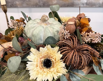 May include: A decorative centerpiece featuring a pale green pumpkin, a woven twig pumpkin, and a large cream-colored daisy. The arrangement includes eucalyptus leaves, brown acorns, dried poppy pods, and small white floral sprigs, creating a textured, multi-layered botanical display.