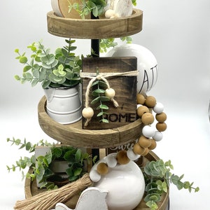 May include: A three-tiered wooden tray stand with a black metal frame. The top tier has a wooden slice, a white ceramic ornament, and greenery. The middle tier has a white ceramic pitcher, a wooden sign that says "Home", and wooden beads. The bottom tier has a white ceramic bird, a wooden sign that says "Sweet", and wooden beads.