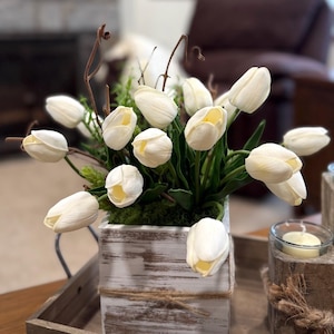 May include: A display of white tulips with yellow centres, presented in a distressed white wooden box tied with twine. The arrangement includes green foliage and twigs. A candle in a glass holder is visible on the right, enhancing the natural theme.