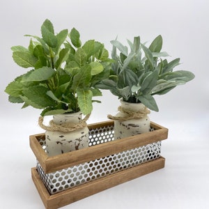 May include: A decorative wooden tray with two small, distressed white vases, each holding faux greenery. The tray has a perforated metal band and is tied with rope. The greenery includes green and gray-green leaves.