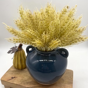 May include: A dark blue ceramic vase with two handles holds a bundle of dried wheat. A small yellow pumpkin with a metal leaf accent sits on a wooden tray. The arrangement is set against a white background.