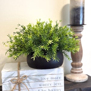 May include: A decorative arrangement featuring a black bowl filled with green foliage, resting atop stacked white books tied with twine. The books display the text "live more worry less and be happy". A tall candle and wooden candle holder are in the background.