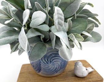 May include: A small blue and white ceramic pot with a swirling pattern containing artificial sage-like green leaves, placed on a rectangular wooden riser next to a small white ceramic bird figurine against a plain white background.