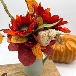 Puede incluir: Un arreglo floral de temática otoñal en una jarra verde salvia. El arreglo incluye girasoles rojos, hojas naranjas y rojas y una pequeña calabaza. Una calabaza y piñas están en el fondo, todo sobre una superficie de madera.