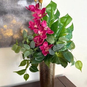以下が含まれることがあります： テクスチャ加工された表面の背の高い金色の花瓶には、鮮やかなフラワーアレンジメントが飾られています。アレンジメントは、明るいピンクの蘭、大きな緑のモンステラの葉、およびさまざまな緑の葉で構成されています。花瓶は濃い木製の表面に置かれています。