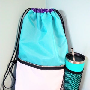May include: A turquoise and white drawstring backpack with a black zippered pocket and a black mesh cup holder. The cup holder has a turquoise travel mug with a silver lid and a stainless steel straw.
