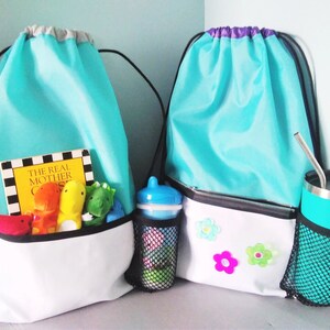 May include: Two turquoise drawstring bags with white accents. One bag has a mesh pocket with a blue sippy cup and a book titled "The Real Mother Goose". The other bag has a mesh pocket with a turquoise travel mug and three colorful flower appliques.
