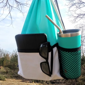 May include: A turquoise drawstring bag with a white bottom and black trim. The bag has a black mesh pocket on the side and a black tablet and sunglasses inside. A turquoise travel mug with a black lid and a stainless steel straw is in the mesh pocket.