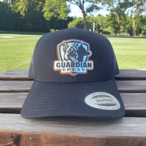 May include: Black and silver trucker hat with a brown patch featuring a German Shepherd silhouette and the text "GUARDIAN EPOXY". The hat has a mesh back and a snap closure.