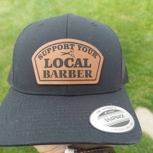 May include: Black trucker hat with a brown leather patch that says "Support Your Local Barber" with a pair of scissors graphic.