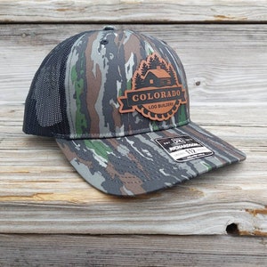 May include: A camouflage trucker hat with a brown leather patch that says "Colorado Log Builders" with a small cabin image and pine trees.