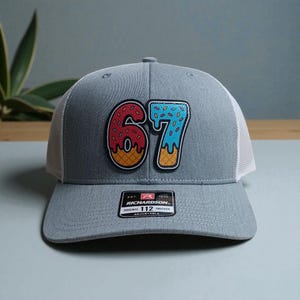 May include: Gray trucker hat with a white mesh back and a curved brim. The front features a colorful patch with the number "67" designed to look like donuts. The hat includes a "RICHARDSON" tag.