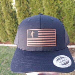 May include: A black and brown trucker hat with a copper-colored American flag featuring a lineman silhouette. The hat has a black mesh back and a black snap closure. The hat has a "Snapback" label on the back.