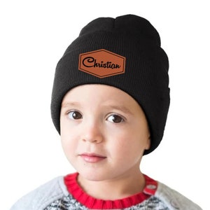 May include: Black beanie with a brown leather patch that reads "Christian" in a cursive font. The beanie is worn by a young child. The child is wearing a gray and red sweater.