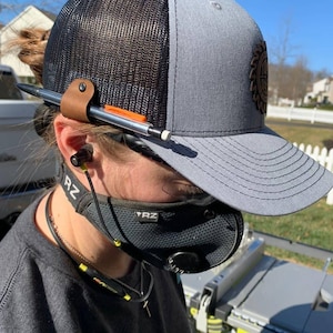 May include: A person wearing a gray and black baseball cap with a logo, a black face mask with the text "YRZ", and black earbuds. The person is wearing a gray shirt and has a necklace with a yellow pendant.