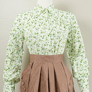 Könnte beinhalten: Ein Outfit im Vintage-Stil mit einer weißen Bluse mit grünem Blumenmuster und einem braunen Faltenrock. Die Bluse hat lange Ärmel und eine Knopfleiste. Der Rock hat eine hohe Taille und Knöpfe.