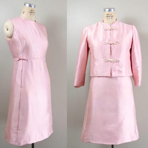Puede incluir: Un vestido vintage rosa claro y un conjunto de chaqueta a juego. El vestido sin mangas tiene un corpiño ajustado y una falda a la rodilla. La chaqueta presenta un cuello mandarín y cierres de nudo decorativos. Ambas piezas se exhiben en maniquíes.