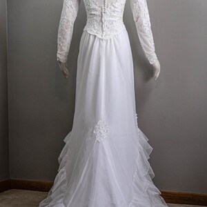 May include: A white wedding dress with long sleeves and a high neckline. The dress has a lace bodice and a flowing skirt with ruffles.