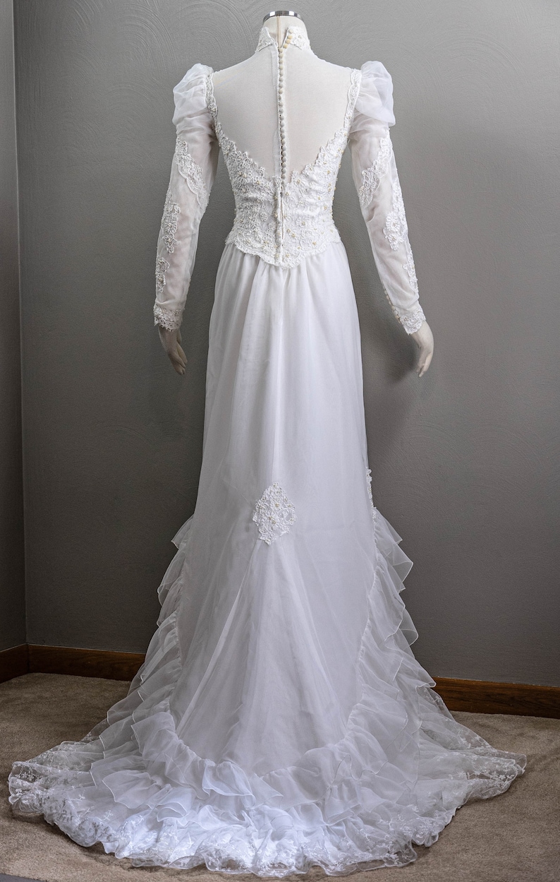 May include: A white wedding dress with long sleeves and a high neckline. The dress has a lace bodice and a flowing skirt with ruffles.