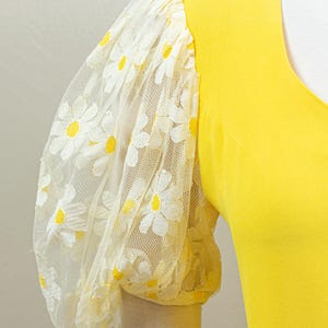 May include: A bright yellow top with a V-neck and a sheer, puffy sleeve. The sleeve is made of white lace with a daisy pattern featuring yellow centers. The sleeve cuff is a solid yellow.