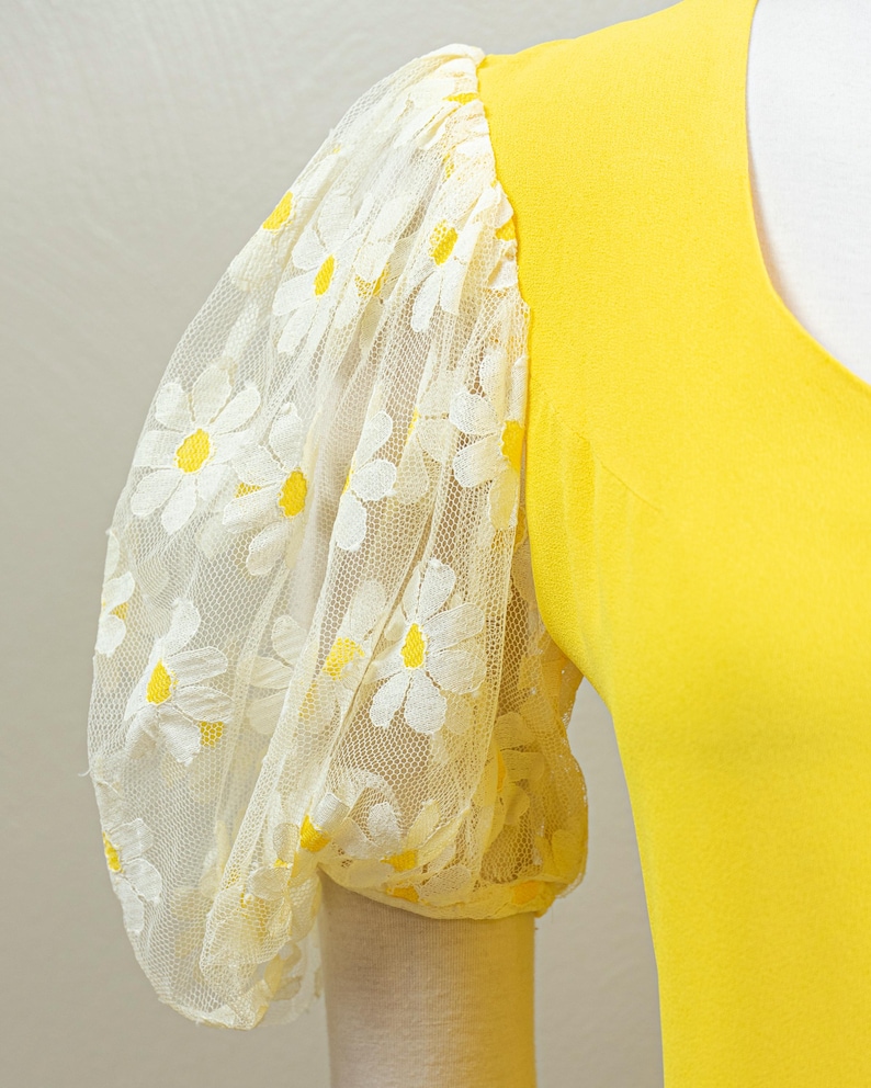May include: A bright yellow top with a V-neck and a sheer, puffy sleeve. The sleeve is made of white lace with a daisy pattern featuring yellow centers. The sleeve cuff is a solid yellow.