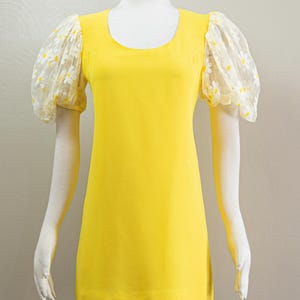 May include: A bright yellow dress with a scoop neckline and short sleeves. The sleeves are made of sheer fabric and feature a white daisy pattern. The dress is displayed on a mannequin.