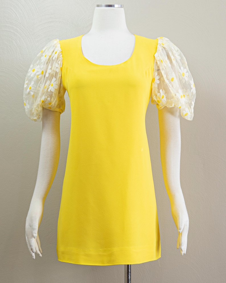 May include: A bright yellow dress with a scoop neckline and short sleeves. The sleeves are made of sheer fabric and feature a white daisy pattern. The dress is displayed on a mannequin.