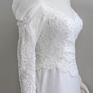 May include: A white wedding dress with long sleeves and a high neckline. The dress is made of lace and tulle and features a fitted bodice and a flowing skirt.