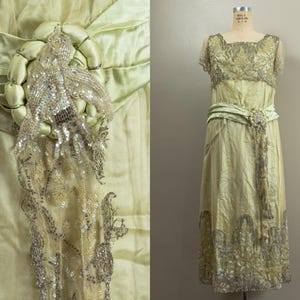 May include: A vintage pale green dress with intricate silver beading and embellishments. The dress features a square neckline, short sleeves, and a flowing skirt. A matching sash cinches the waist, and a decorative beaded element adorns the bodice. The dress is displayed on a mannequin.