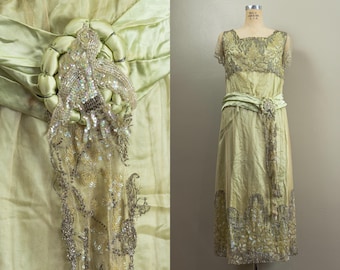 May include: A vintage pale green dress with intricate silver beading and embellishments. The dress features a square neckline, short sleeves, and a flowing skirt. A matching sash cinches the waist, and a decorative beaded element adorns the bodice. The dress is displayed on a mannequin.