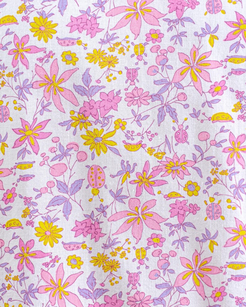 Puede incluir: Una tela blanca con un estampado floral repetitivo. El dise&ntilde;o presenta flores rosas, amarillas y moradas, junto con peque&ntilde;as ilustraciones de mariquitas. Las flores var&iacute;an en tama&ntilde;o y forma, creando una impresi&oacute;n caprichosa y colorida.