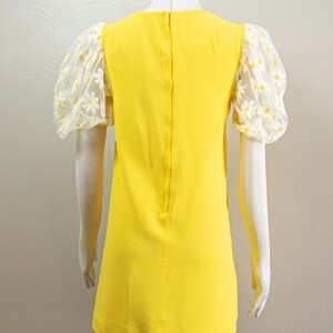 May include: A bright yellow dress with sheer, daisy-patterned sleeves. The dress has a simple, A-line silhouette and a high neckline. The sleeves are puffy and made of a white mesh fabric with yellow and white daisy embroidery. The dress is displayed on a mannequin.