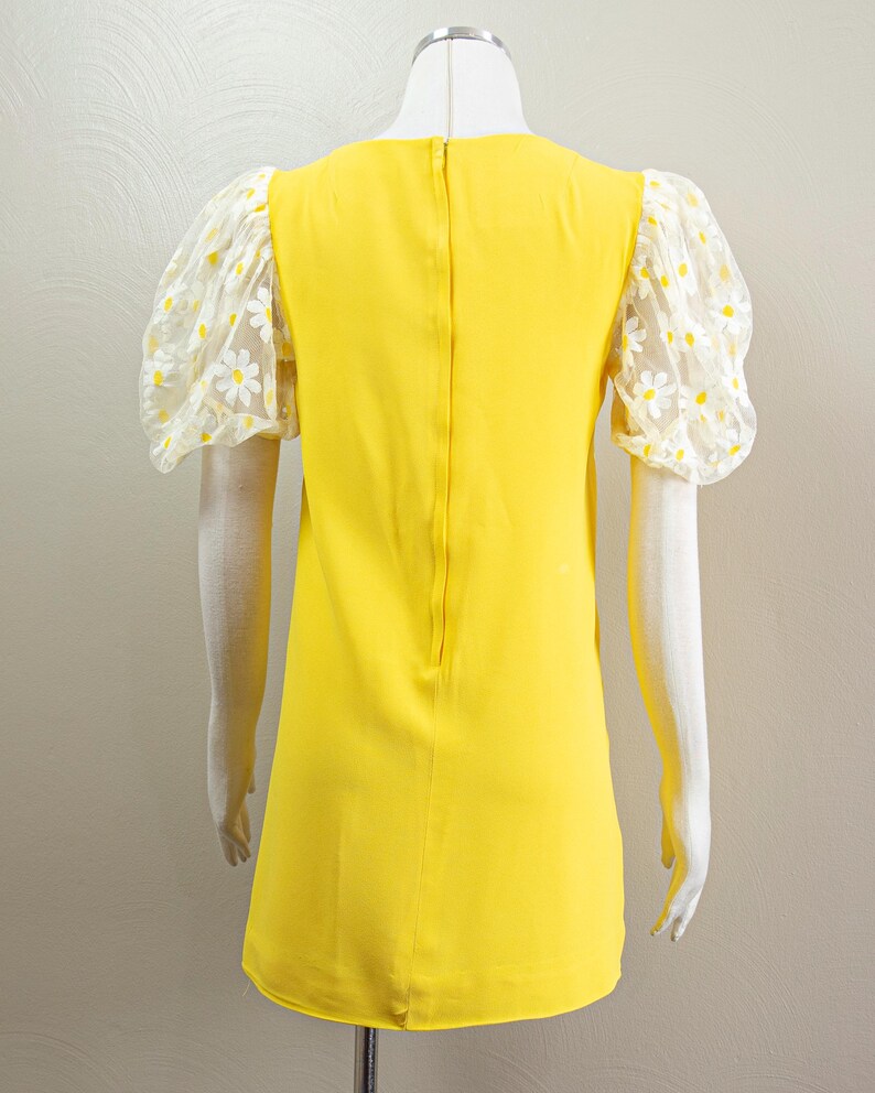 May include: A bright yellow dress with sheer, daisy-patterned sleeves. The dress has a simple, A-line silhouette and a high neckline. The sleeves are puffy and made of a white mesh fabric with yellow and white daisy embroidery. The dress is displayed on a mannequin.