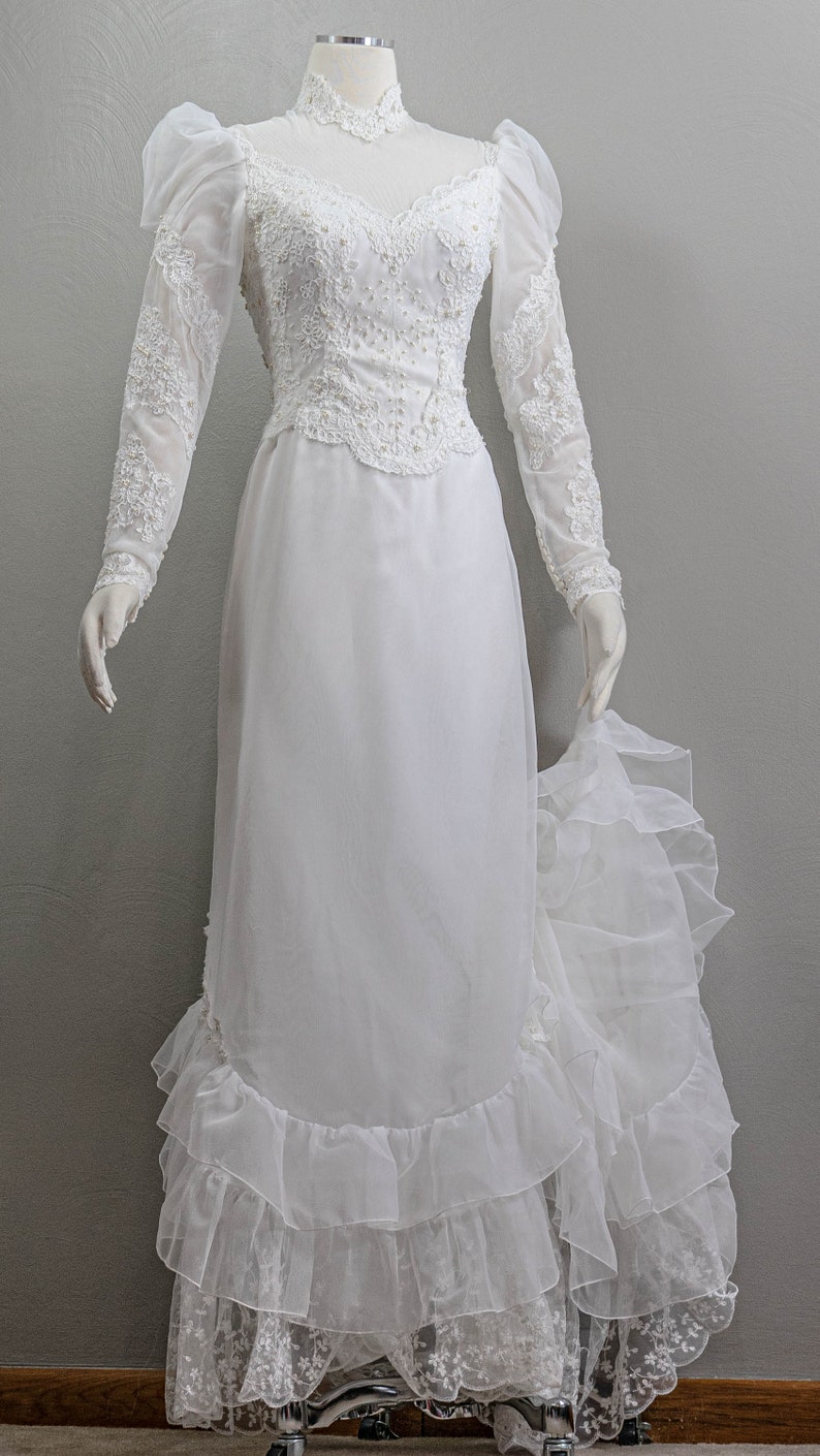 May include: A white wedding dress with a high neckline, long sleeves, and a lace bodice. The skirt is made of layers of tulle and has a ruffled hem.