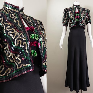 Puede incluir: Una chaqueta de bolero negra con lentejuelas de color verde, dorado y rojo. La chaqueta se lleva sobre un vestido negro con un corpiño ajustado y una falda fluida.