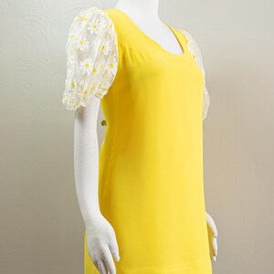 May include: A bright yellow dress with a scoop neckline and short, sheer sleeves. The sleeves are adorned with a white daisy pattern. The dress is displayed on a mannequin.