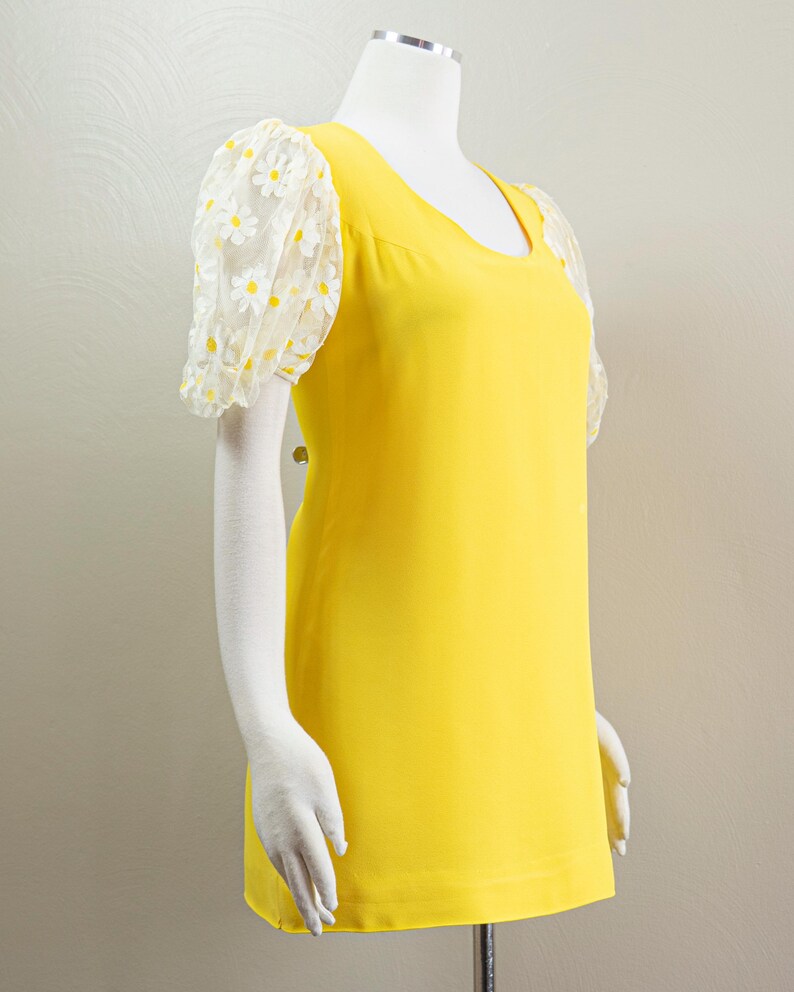 May include: A bright yellow dress with a scoop neckline and short, sheer sleeves. The sleeves are adorned with a white daisy pattern. The dress is displayed on a mannequin.