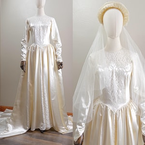 Puede incluir: Un vestido de novia vintage de color marfil con mangas largas y una falda amplia. El corpiño presenta intrincados detalles de encaje y el vestido tiene una larga cola. Un velo y un tocado a juego completan el conjunto nupcial, exhibido en un maniquí.