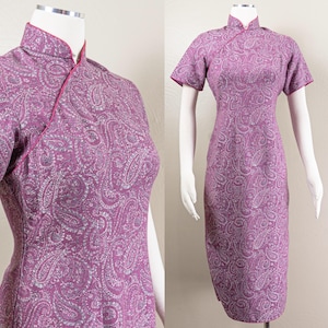 May include: A vintage purple dress with a paisley pattern. The dress has short sleeves, a mandarin collar, and a fitted shape. The edges are trimmed with pink piping. The dress is displayed on a mannequin.