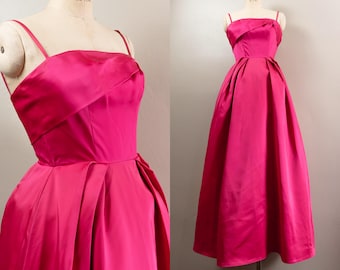 May include: A vibrant fuchsia-colored formal gown. The dress features a fitted bodice with a draped neckline and thin spaghetti straps. The full skirt is pleated, creating a classic silhouette. The dress is displayed on a mannequin.