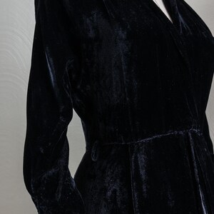 K&ouml;nnte beinhalten: Ein schwarzes Samtkleid mit V-Ausschnitt und langen &Auml;rmeln. Das Kleid hat eine taillierte Taille und einen flie&szlig;enden Rock.