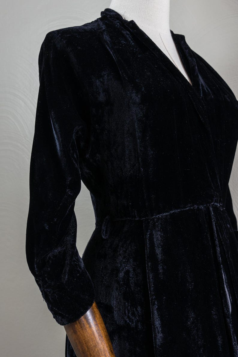 K&ouml;nnte beinhalten: Ein schwarzes Samtkleid mit V-Ausschnitt und langen &Auml;rmeln. Das Kleid hat eine taillierte Taille und einen flie&szlig;enden Rock.